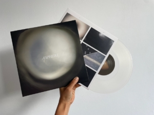 Een hand houdt de LP ‘Asphere’ vast met hoes, transparante vinylplaat en bijhorende insert met foto’s.