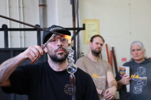 Glasblazer tilt een kettingvormig, transparant glas omhoog; aan het uiteinde hangt een gloeiende druppel, toeschouwers kijken mee op de achtergrond.