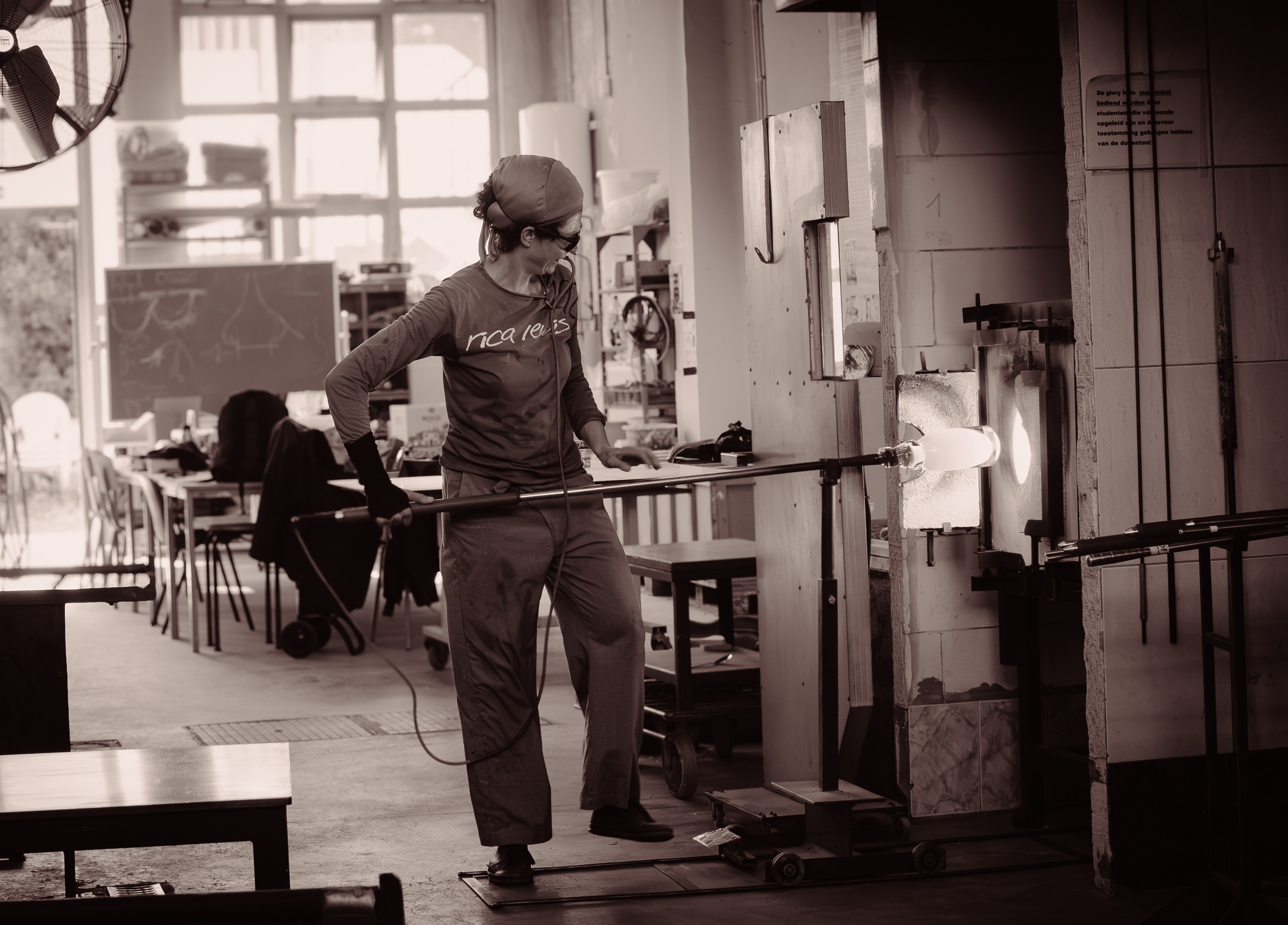 Een leerling glas werkt aan een blaaspijp bij de glasoven in het atelier. Het gloeiende glas wordt voor de ovenopening verhit.